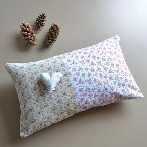 以下が含まれることがあります： 花柄の四角い枕。枕の一方は薄い茶色で、小さな濃い茶色の花が咲いています。もう一方は白で、小さなピンクの花が咲いています。小さな白いハート型の枕が枕の上に置かれています。