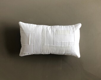 May include: White rectangular pillow with a patchwork design. The pillow is made of white fabric with different sized squares sewn together.