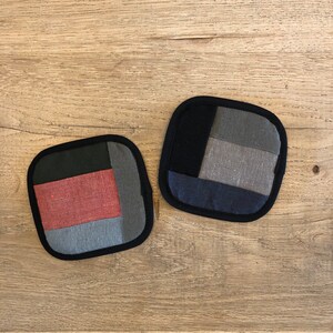 以下が含まれることがあります： パッチワークのデザインが施された正方形のキルト鍋つかみが2つ。1つの鍋つかみは黒、グレー、ブルー。もう1つの鍋つかみは黒、グレー、赤、ブルーです。