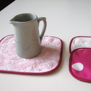 May include: Two pink and white pot holders with a burgundy trim. One pot holder has a gray ceramic pitcher on top of it.