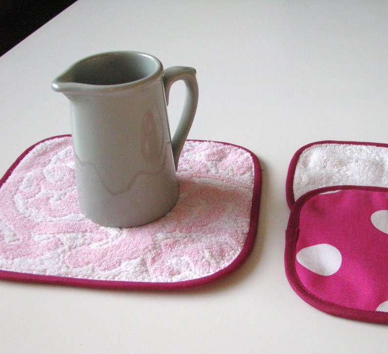 May include: Two pink and white pot holders with a burgundy trim. One pot holder has a gray ceramic pitcher on top of it.