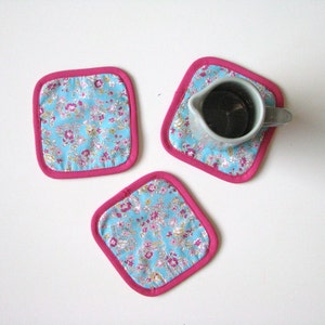 May include: Three pink-trimmed blue floral fabric coasters. One coaster has a small gray ceramic pitcher on top.