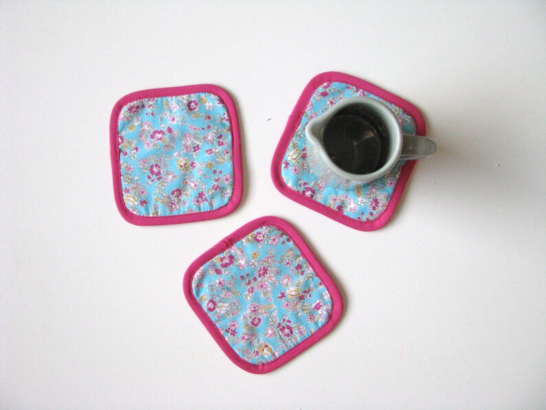May include: Three pink-trimmed blue floral fabric coasters. One coaster has a small gray ceramic pitcher on top.
