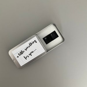 May include: A small silver tin with a white lined paper inside that says "a little something for you..." The tin is open and reveals two black keys with the letters "H" and "I" on them.