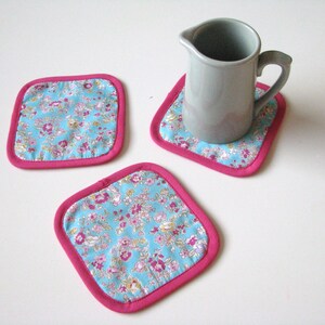 May include: Three pink and blue floral fabric coasters with a gray ceramic pitcher on one of the coasters.