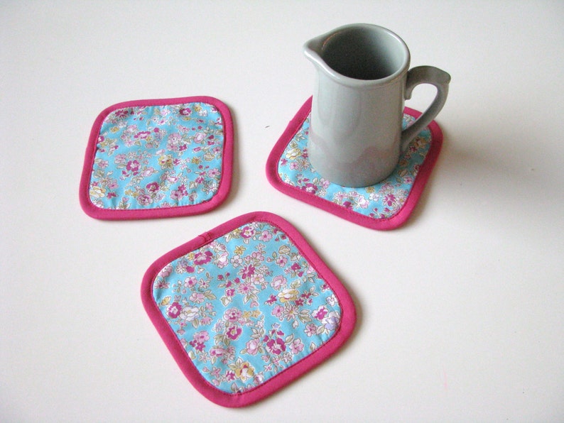 May include: Three pink and blue floral fabric coasters with a gray ceramic pitcher on one of the coasters.