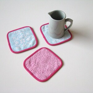 May include: Three square fabric coasters with pink and blue trim. Two coasters have a white floral pattern and one is solid pink.