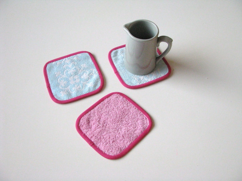 May include: Three square fabric coasters with pink and blue trim. Two coasters have a white floral pattern and one is solid pink.