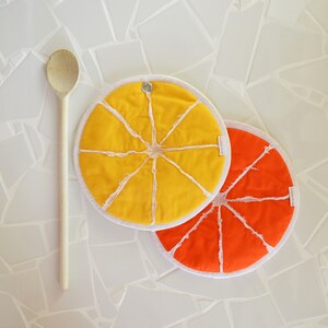 Peut inclure: Deux dessous de plat ronds orange et jaune avec une bordure blanche et un motif cousu ressemblant à une tranche d'agrume. Les dessous de plat sont sur une surface carrelée blanche avec une cuillère en bois.