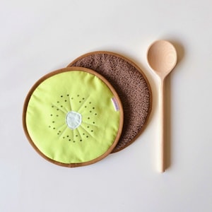 Könnte beinhalten: Zwei braune und grüne Topflappen in Form von Kiwis. Ein Topflappen ist aus braunem Stoff mit einem Kiwi-Motiv auf der Oberseite. Der andere Topflappen ist aus braunem Stoff mit einem Kiwi-Motiv auf der Oberseite. Ein Holzlöffel liegt neben den Topflappen.