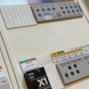 May include: A white electrical panel with a black box labeled "X1" and a white control panel with buttons and labels for different rooms, including "Windfang", "Gäste Bad", "Küche", "Wohnen", and "Essen".