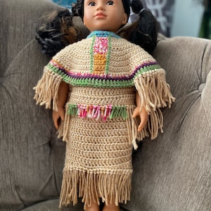 Puede incluir: Una muñeca con un atuendo de ganchillo con flecos. El atuendo es de color canela con rayas de colores y un ribete rosa, verde y naranja. La muñeca tiene el pelo largo y negro y los ojos marrones.