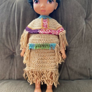 Puede incluir: Un vestido de crochet marrón con flecos y un poncho de crochet de colores en una muñeca con cabello y ojos marrones.