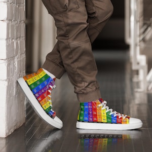 Peut inclure: Une paire de baskets montantes en toile blanche avec un imprimé tableau périodique arc-en-ciel. Les baskets sont portées par une personne portant un pantalon marron.