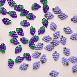 May include: A selection of purple grape-shaped embellishments with green leaves and red stems. These decorative pieces are scattered on a light pink surface, ideal for crafting or scrapbooking projects.