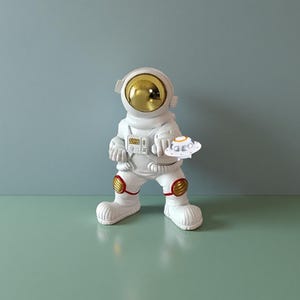 May include: A white astronaut figurine with a gold visor and red and gold accents on the knees. The astronaut holds a small, white UFO. The figurine is standing on a green surface.