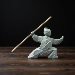 Op de afbeelding: Een grijze stenen sculptuur van een persoon in een vechtsportpose, die een houten staf vasthoudt. De figuur knielt met één been uitgestrekt en armen uitgestrekt. De sculptuur staat op een donker houten oppervlak met een zwarte achtergrond.
