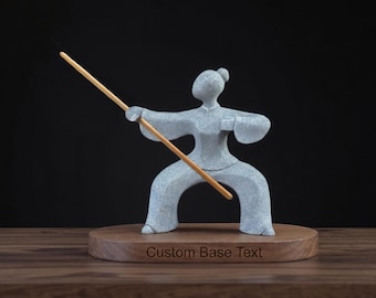 May include: A light blue, textured figurine of a person in a martial arts stance holding a long, thin wooden staff. The figure is mounted on an oval wooden base with the engraved text "Custom Base Text" centred on the front edge of the wood.