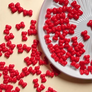 May include: Numerous small, glossy red bow-shaped decorations are scattered on a pale yellow surface and a white plate. The bows are evenly spaced, forming an attractive pattern. These decorative items could be used for crafts or other creative projects.