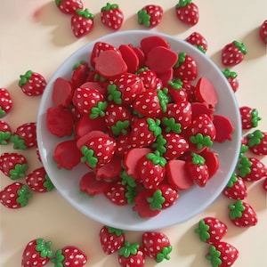 Op de afbeelding: Een verzameling rode aardbeivormige versieringen met groene steeltjes en witte spikkels, sommige zijn heel en sommige zijn gesneden. De aardbeien zijn verspreid rond en in een witte kom.