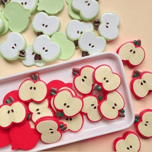May include: A collection of apple-shaped embellishments in red, green, and white. The red apples are cut in half, revealing a cream-colored interior with black seeds. Green and white apple halves are also present, along with whole green apple shapes.