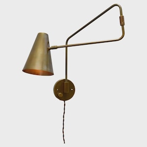 Op de afbeelding: Een wandlamp met een conische koperen kap en een verstelbare arm. Het armatuur heeft een ronde muurplaat met een kleine bedieningsknop en een bruin gedraaid stoffen snoer dat vanaf de basis naar beneden loopt, uitgevoerd in een metallic koperkleur.