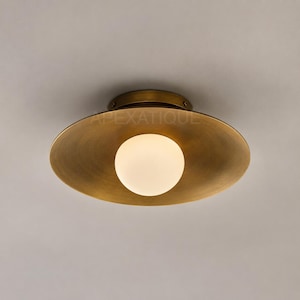May include: A flush mount ceiling light fitting featuring a wide, circular, brushed brass metal shade with a warm metallic finish. A single, spherical, frosted white glass globe bulb is centred within the shade. The text APEXATIQUE is visible.