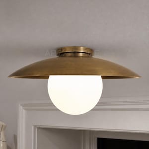 May include: A flush mount ceiling light fitting featuring a wide, shallow, brushed brass metal shade with a smooth, curved profile. A large, spherical, opaque white glass globe bulb is centred beneath the metal shade, providing illumination.