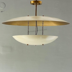 Op de afbeelding: Een hanglamp in mid-century modern stijl met een messing afwerking en twee off-white, schotelvormige kappen. De armatuur hangt aan een messing staaf en ketting. De bovenste kap is groter dan de onderste kap.