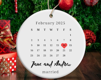 May include: A white ceramic Christmas ornament with a gold string for hanging. The ornament features a calendar for February 2025, with the 14th highlighted with a red heart. The names "Jane and Andrew" and the word "married" are also printed on the ornament.
