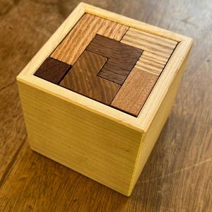 May include: A wooden box puzzle with a variety of wood tones, including light brown, dark brown, and reddish brown. The puzzle pieces are arranged in a geometric pattern inside the box.
