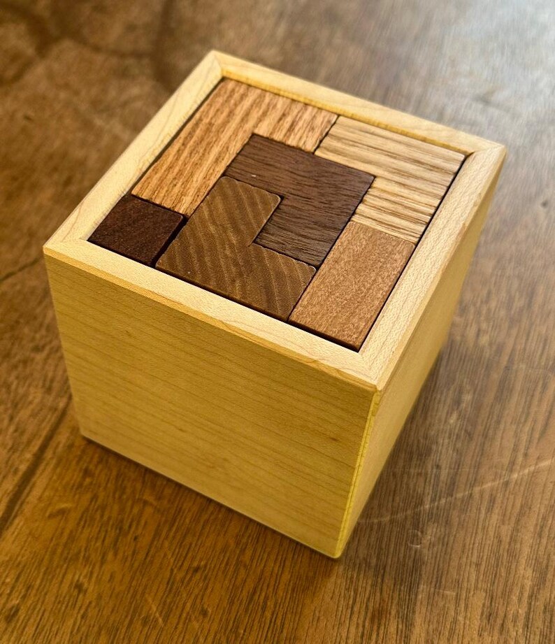 May include: A wooden box puzzle with a variety of wood tones, including light brown, dark brown, and reddish brown. The puzzle pieces are arranged in a geometric pattern inside the box.