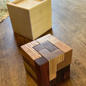 May include: A wooden puzzle cube made of various wood types, including light brown, dark brown, and reddish brown. The cube is assembled with interlocking pieces, creating a complex geometric pattern.