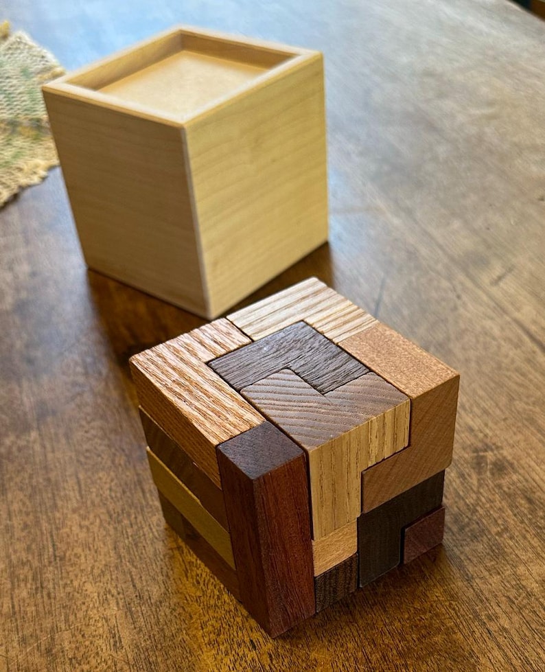 May include: A wooden puzzle cube made of various wood types, including light brown, dark brown, and reddish brown. The cube is assembled with interlocking pieces, creating a complex geometric pattern.