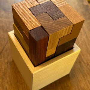 May include: A wooden puzzle box with a cube-shaped puzzle inside. The puzzle is made of different colored wood pieces that fit together to form a larger cube. The box is made of light wood and has a simple design.