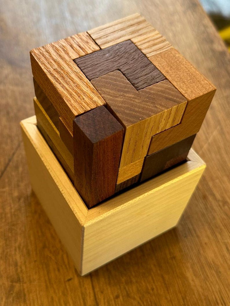 May include: A wooden puzzle box with a cube-shaped puzzle inside. The puzzle is made of different colored wood pieces that fit together to form a larger cube. The box is made of light wood and has a simple design.
