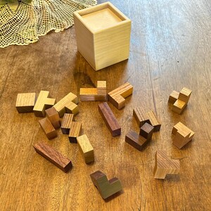 May include: A wooden box with a lid, containing a set of wooden puzzle pieces. The pieces are in various shapes and sizes, and are made of different types of wood. The pieces are scattered on a wooden table.