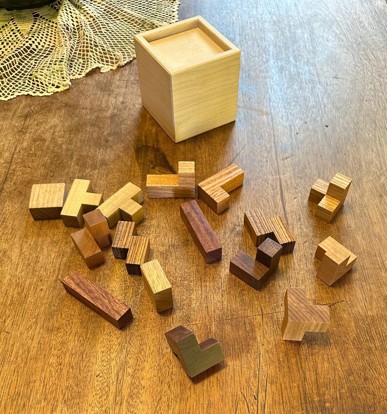 May include: A wooden box with a lid, containing a set of wooden puzzle pieces. The pieces are in various shapes and sizes, and are made of different types of wood. The pieces are scattered on a wooden table.
