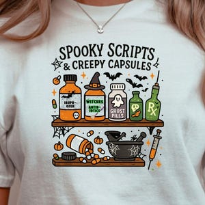 May include: White t-shirt with a Halloween-themed graphic. The design features a shelf with potion bottles, a mortar and pestle, and the text "Spooky Scripts & Creepy Capsules". The design is in orange, green, black, and white.
