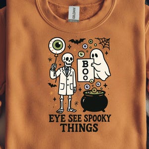 Może przedstawiać: Pomarańczowa bluza z grafiką o tematyce Halloween. Projekt przedstawia szkielet trzymający balon z okiem, ducha, kocioł i napis "EYE SEE SPOOKY THINGS". Nietoperze i gwiazdy dopełniają projekt.