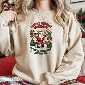 Puede incluir: Sudadera beige con un gráfico festivo. El diseño presenta a Papá Noel levantando pesas con un elfo, un árbol de Navidad y el texto "SANTA'S HOLIDAY WORKOUT" y "PHYSICAL THERAPIST APPROVED!"