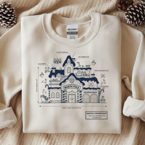 May include: Cream-colored sweatshirt with a detailed blue blueprint design of Santa's Workshop. The graphic includes the words "North Pole," "Reindeer Garage," and other holiday-themed elements. The sweatshirt is made of heavy cotton.