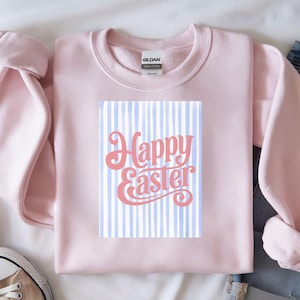 May include: A pale pink sweatshirt featuring the text "Happy Easter" in coral script over a blue and white striped design. The sweatshirt is made of a soft material, ideal for the Easter holiday.