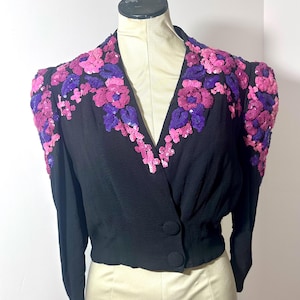 Op de afbeelding: Zwarte jas met lange mouwen, een V-hals en een sluiting met twee knopen. De schouders en halslijn zijn versierd met roze en paarse bloemmotieven van pailletten. De jas wordt op een mannequin gepresenteerd.