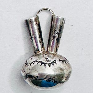 May include: A silver pendant with a rounded base and two cylindrical tubes extending upwards. The base has etched patterns and a small turquoise accent. A small loop is at the top for hanging.