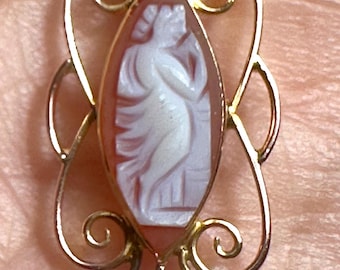 May include: A gold pendant featuring a carved cameo of a classical figure in white against a reddish-brown background. The pendant has an ornate gold filigree frame and a small pearl drop.