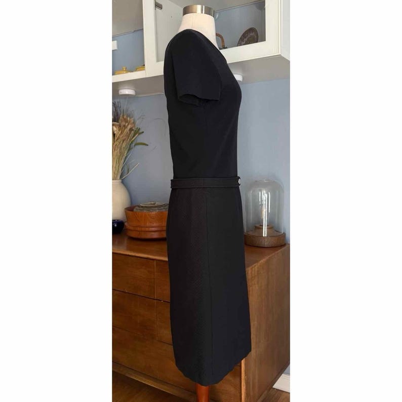 May include: A black dress with short sleeves and a fitted silhouette. The dress features a defined waist with a belt detail. The dress is displayed on a mannequin, showcasing its shape and design. The dress is a classic, elegant garment.