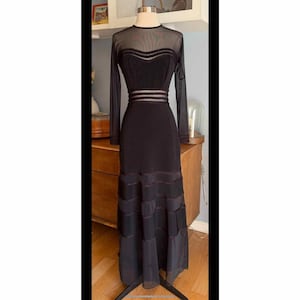 以下が含まれることがあります： 黒のイブニングドレス。長袖とロングスカートが特徴です。ドレスは、透け感のあるメッシュのネックラインと袖、フィットした胴体、ティアードスカートが特徴です。ウエストは水平のシアーパネルでアクセントを添えています。