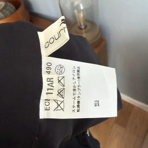 May include: Close-up of a black garment with a white tag. The tag has the brand name "courn" and care symbols. Text on the tag includes "ECE 11AR 490" and Japanese washing instructions. The garment is partially visible.
