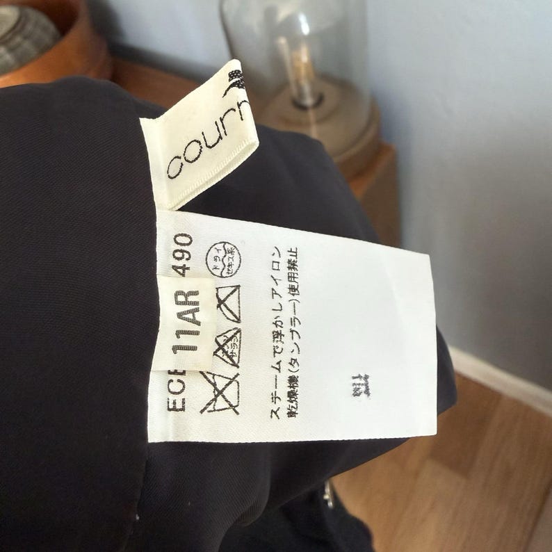 May include: Close-up of a black garment with a white tag. The tag has the brand name "courn" and care symbols. Text on the tag includes "ECE 11AR 490" and Japanese washing instructions. The garment is partially visible.